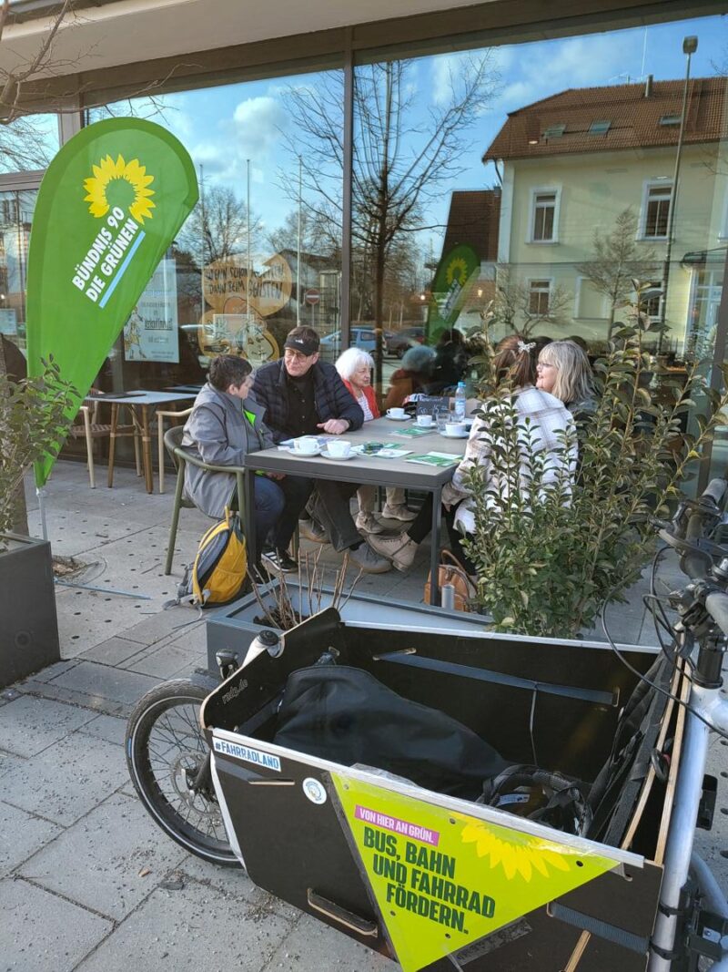 Besetzter Tisch im Aussenbereich des Cafés Aumüller mit Interessierten der GRÜNEN in Feldkirchen und der Gemeindesratskandida:innen. Im Vordergrund eine Beachflag der Bündnis 90/DIE GRÜNEN und ein Lastenrad